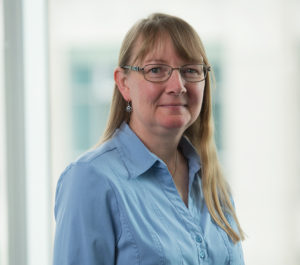 Dr. Clelland is sitting in front of a large window with two floors of a building blurred in the background. She is wearing a buttoned-up blouse.