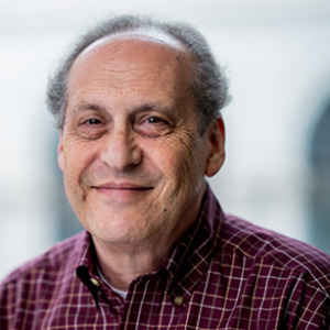Dr. Rubin is sitting in front of a large window with a building blurred in the background. He is wearing a buttoned-up shirt.