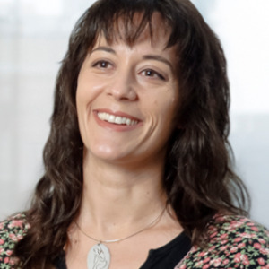 Dr. Maglio is standing in front of a large window with a building blurred in the background. She is wearing a blouse.