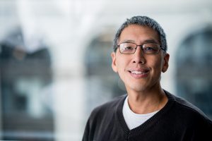 Dr. Ji is sitting in front of a large window with a building blurred in the background. He is wearing a sweater.