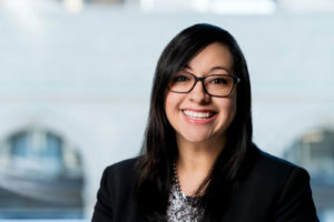 Flor Esquivel sits in front of a large window with buildings blurred in the background. She is wearing a blouse and blazer.