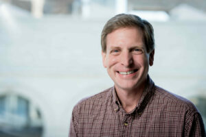 Dr. Finn is standing in front of a large window with a building blurred in the background. He is wearing a buttoned-up shirt.
