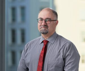 Dr. Sepehry is sitting in front of a large window with a building blurred in the background. He is wearing a buttoned-up shirt and tie.