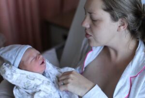 New mother sitting in a hospital bed in pajamas is holding baby, wrapped in a swaddle with a cap.