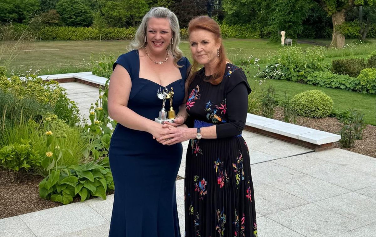 Andrea Carter, dressed in a long formal dress and holding two awards, stands to the left of Sarah Ferguson, also dressed in formal attire.
