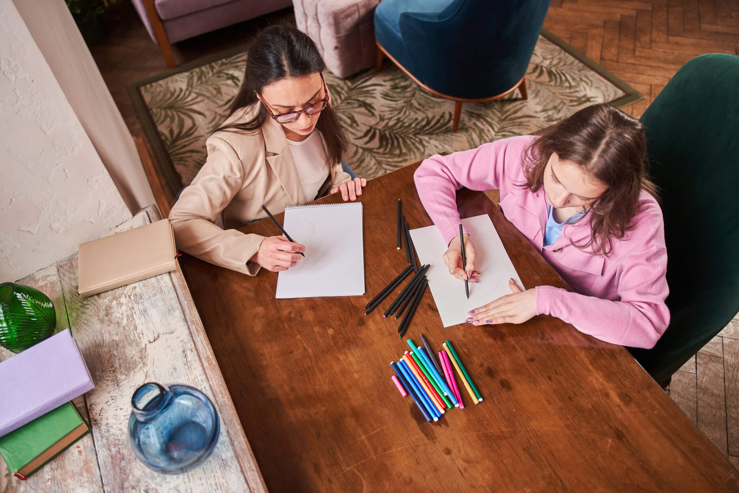 Two women sit at a wooden table, each drawing on white paper with pencils. Colored markers and pencils are spread out between them, suggesting an art therapy session that could inspire creative careers.