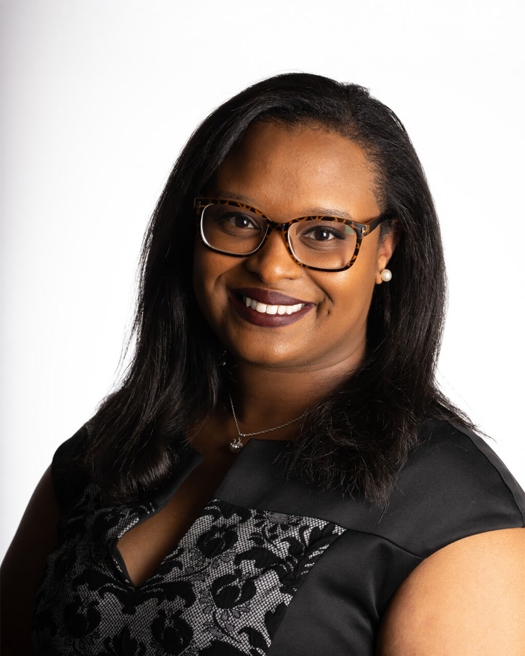 Ashley, a woman with long dark hair and glasses, smiles at the camera in a stylish black lace Poprilo dress, set against a plain white background.