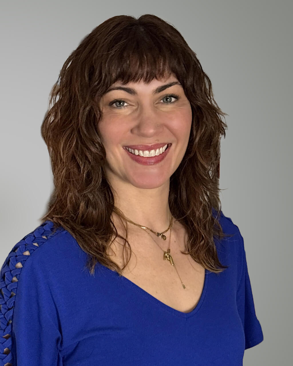 Jennifer Bonino Sobol, a woman with wavy brown hair, wears a blue top and gold necklaces as she smiles at the camera against a neutral Grey background.