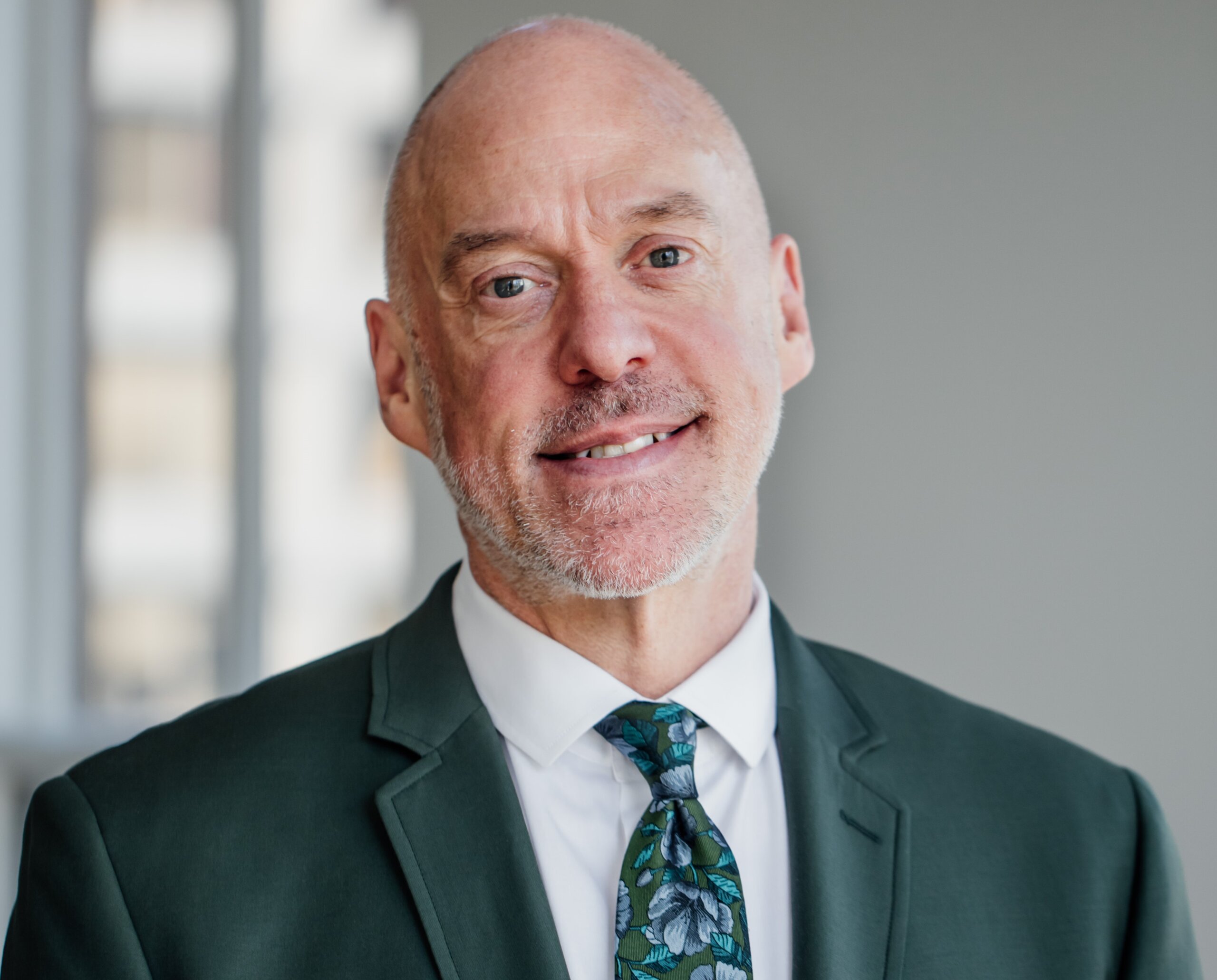 A middle-aged man in a green suit and floral tie smiles at the camera, standing indoors with soft natural light in the background.