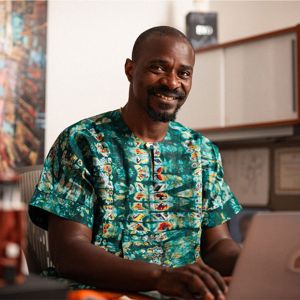A man wearing a green patterned shirt sits at a desk, smiling at the camera, with a laptop in front of him in an office setting, suggesting he may be an Art Therapy Ph.D. professional.