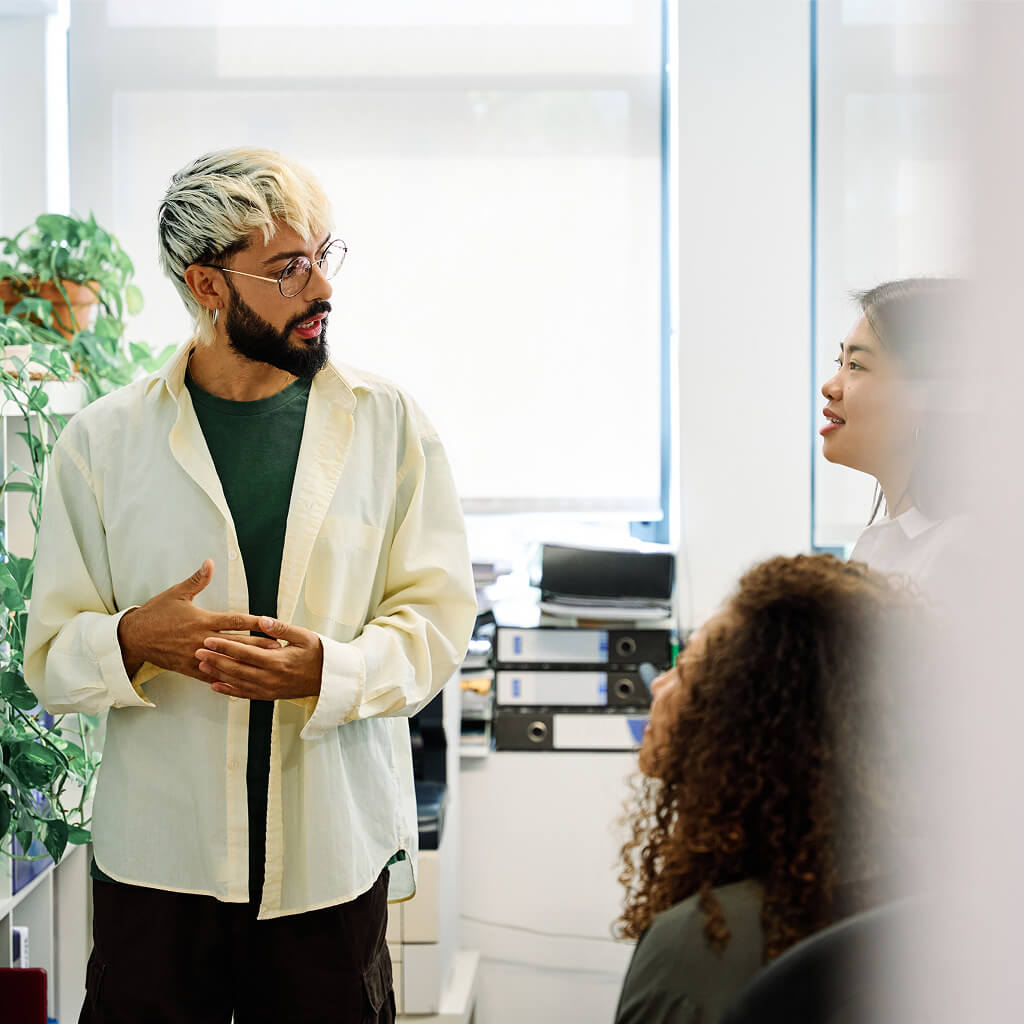 A man with a Ph.D. stands and speaks to two women in an office setting, surrounded by plants, binders, and natural light—discussing the practical applications of Art Therapy.