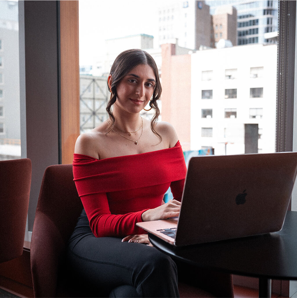 A woman in a red off-shoulder top, possibly an Art Therapy Ph.D. student, sits at a table with an open laptop, looking at the camera. City buildings are visible through the window behind her.