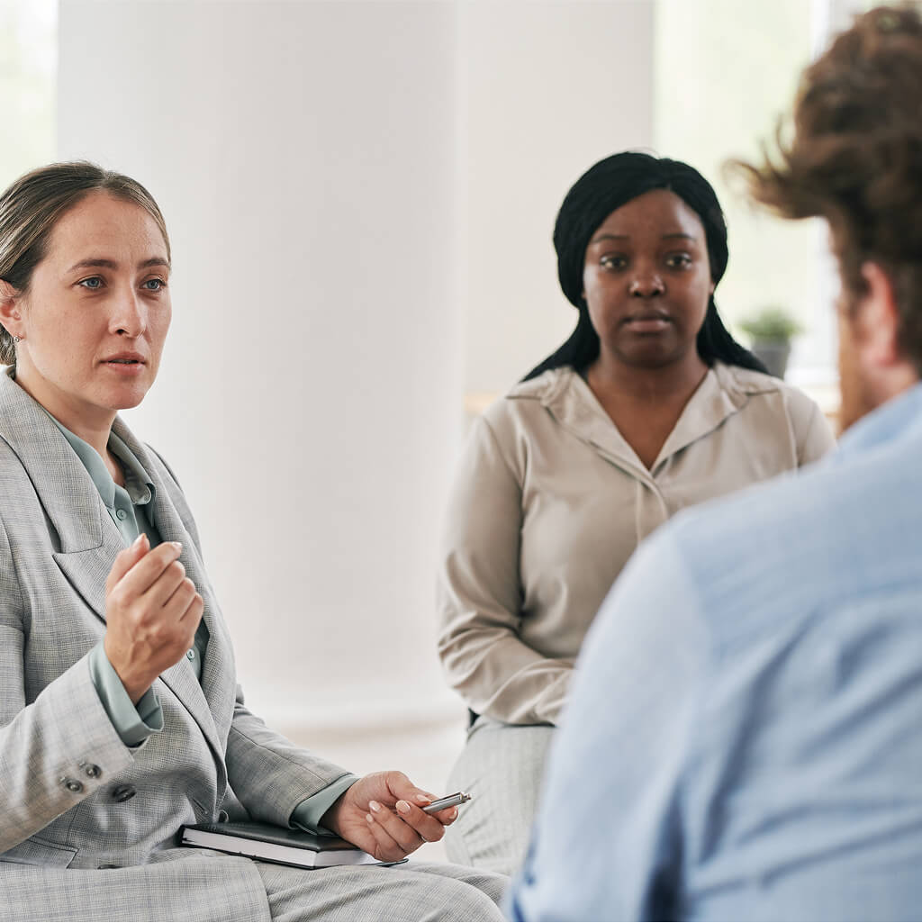 Three people are seated indoors having a serious discussion; one woman, possibly an Art Therapy Ph.D., gestures while holding a notebook, another woman listens intently, and a man is seen from behind.