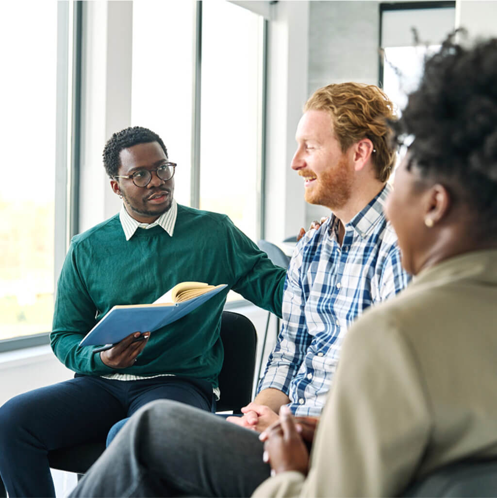 Three people sit together in a modern, bright room; one person with a Ph.D. in Art Therapy holds a notebook, engaging the others in conversation while another smiles.