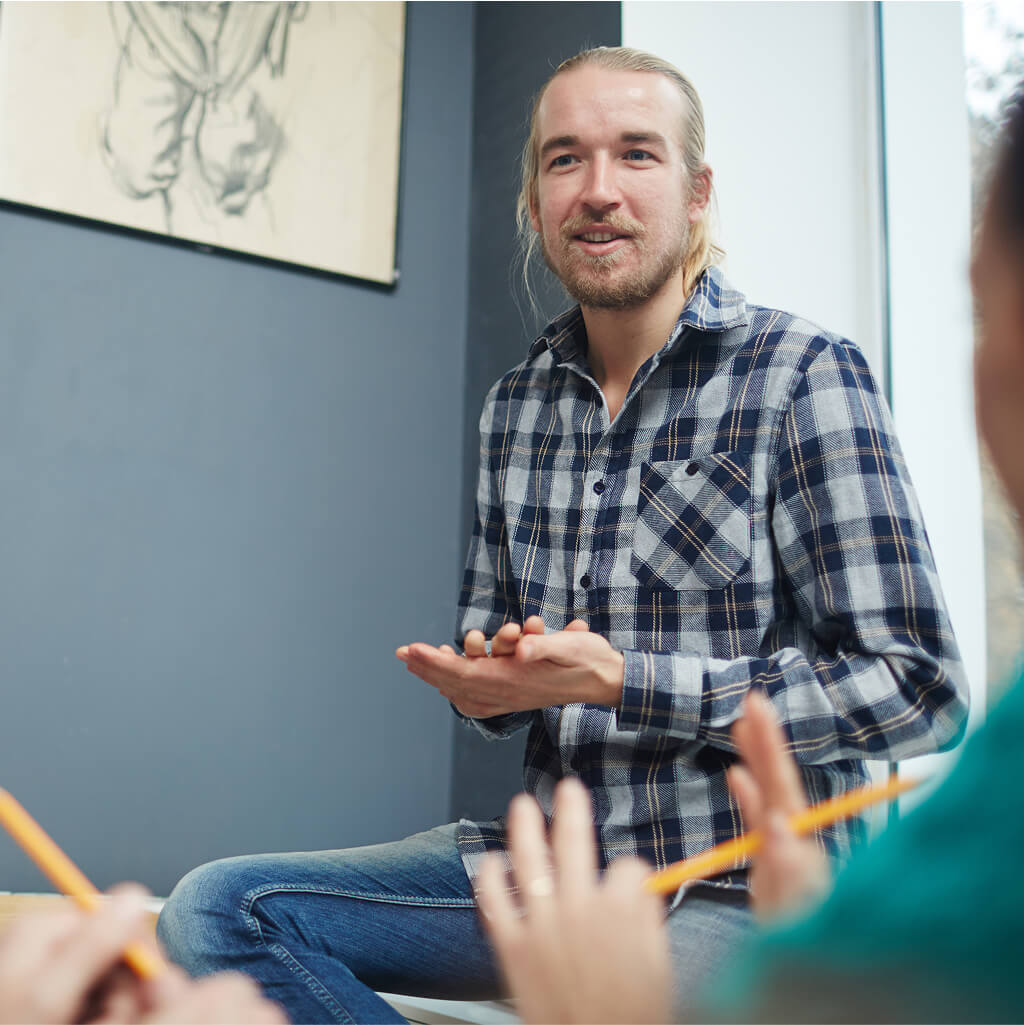 A man in a plaid shirt, possibly an Art Therapy Ph.D., sits and speaks, gesturing with his hands, while two people holding pencils listen in the foreground.