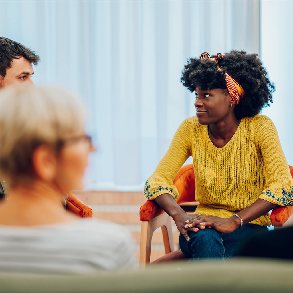 A woman in a yellow sweater sits and listens during a group discussion focused on health and wellness with three other people in a brightly lit room.
