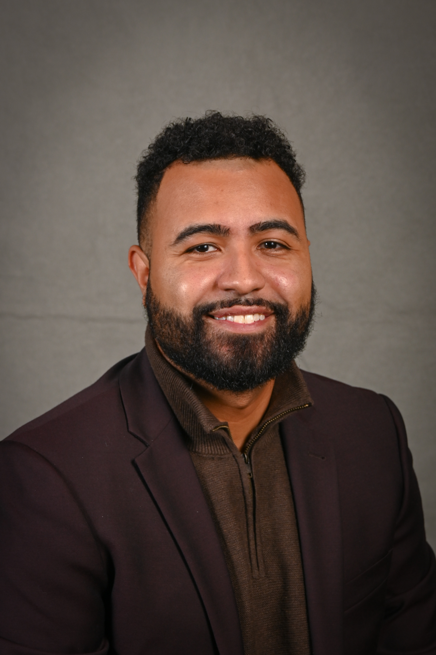 Alexander Perez, a man with a dark curly beard and short hair, wearing a brown blazer and brown shirt, poses and smiles in front of a plain gray background.