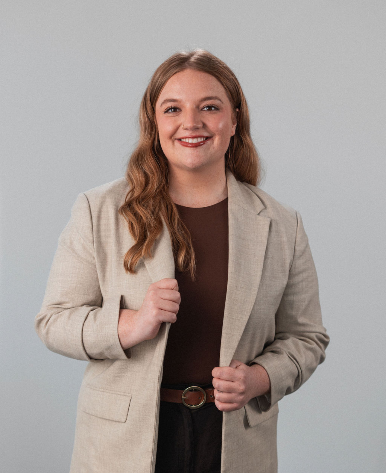 A woman with long light brown hair wearing a beige blazer over a dark top stands against a plain gray background, smiling at the camera.