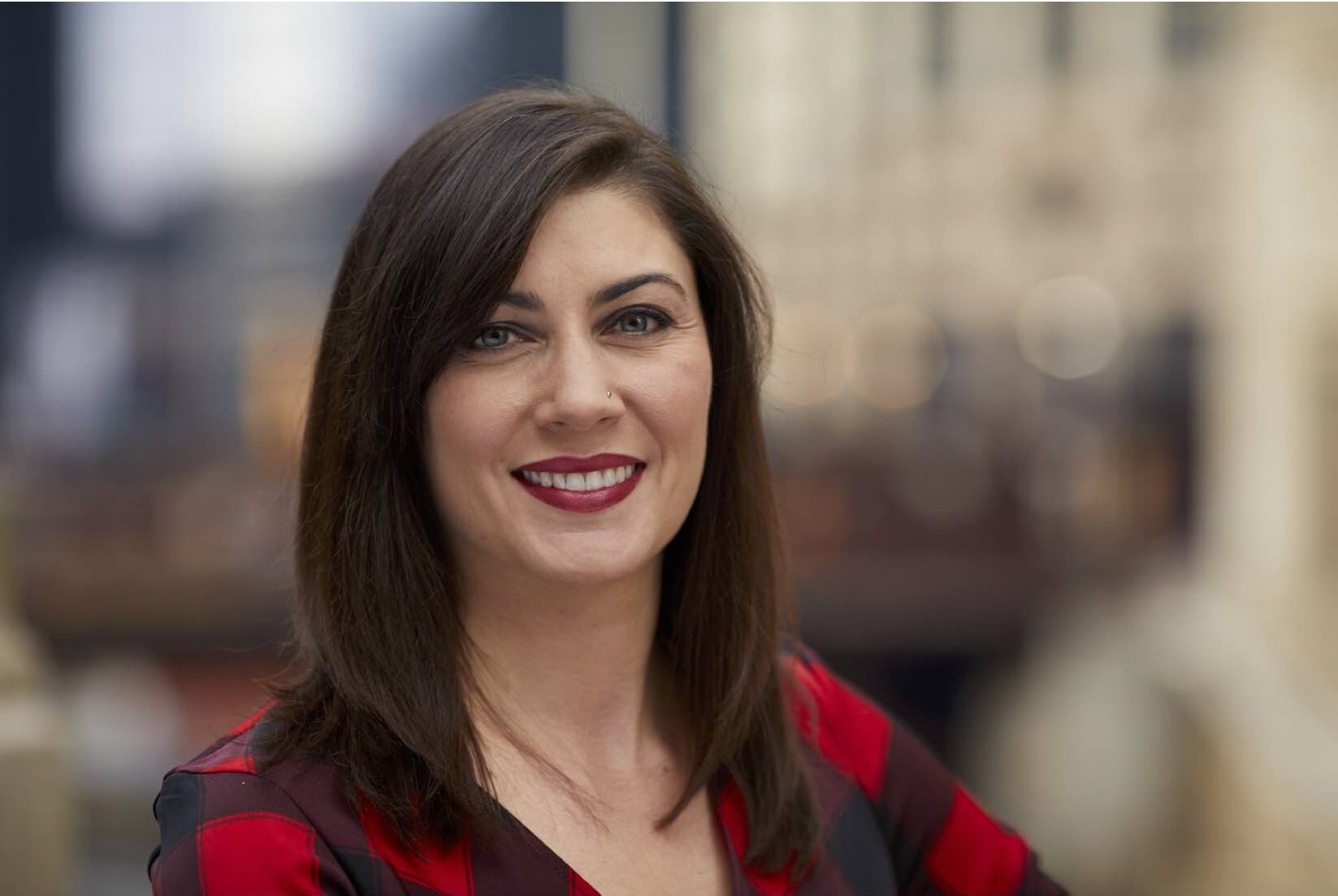 Jennifer, a woman with straight brown hair and a red plaid shirt, smiles at the camera outdoors against a blurred cityscape background.