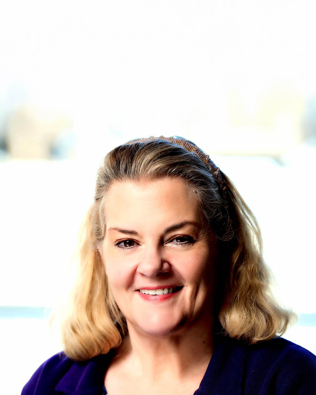 A woman with shoulder-length blonde hair, wearing a dark top, smiles at the camera in a well-lit setting with a blurred background.