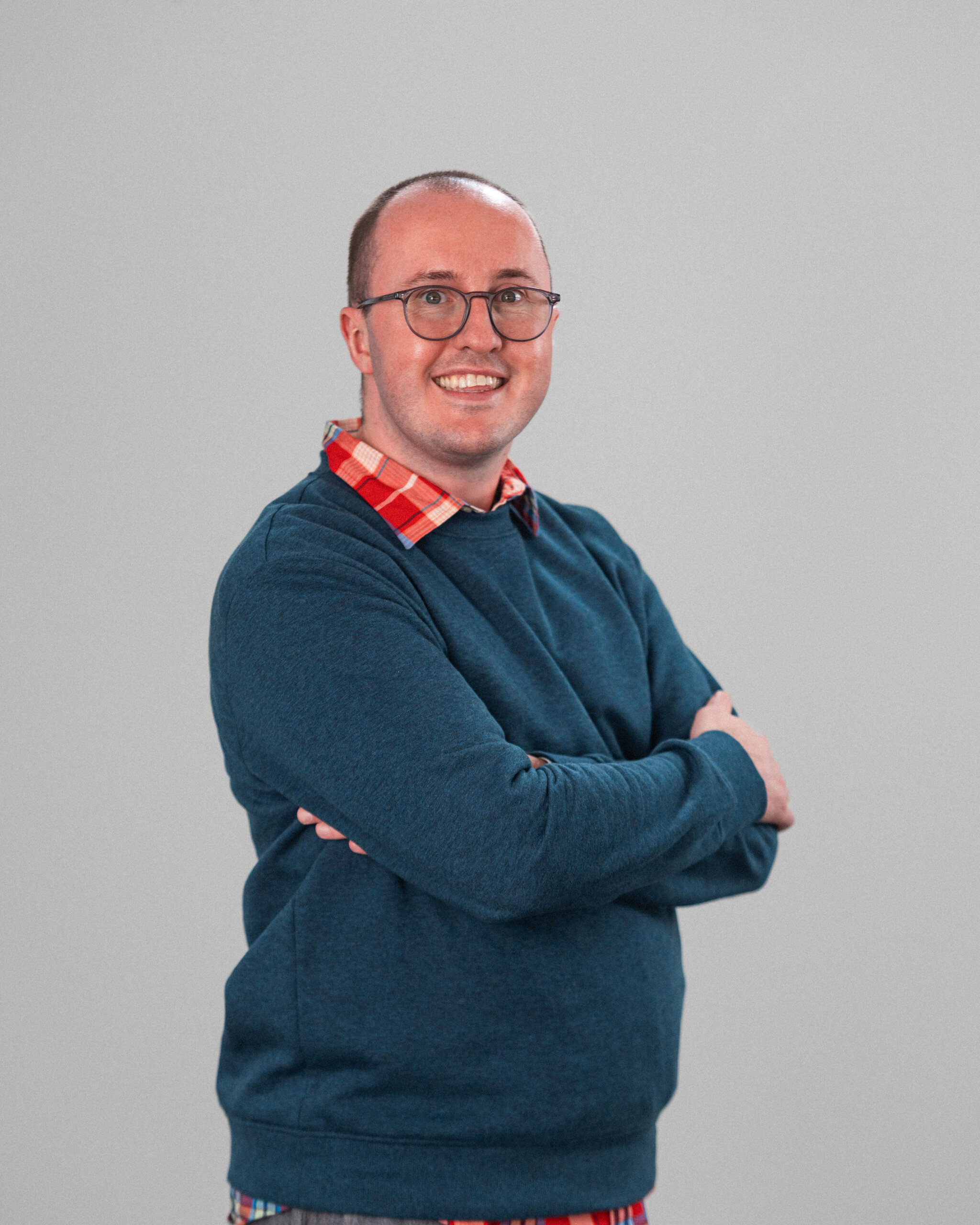 Kyle MacLellan, a person with glasses and short hair, stands with arms crossed and smiles at the camera. Dressed in a blue sweater over a red plaid shirt, he poses confidently against a plain background.