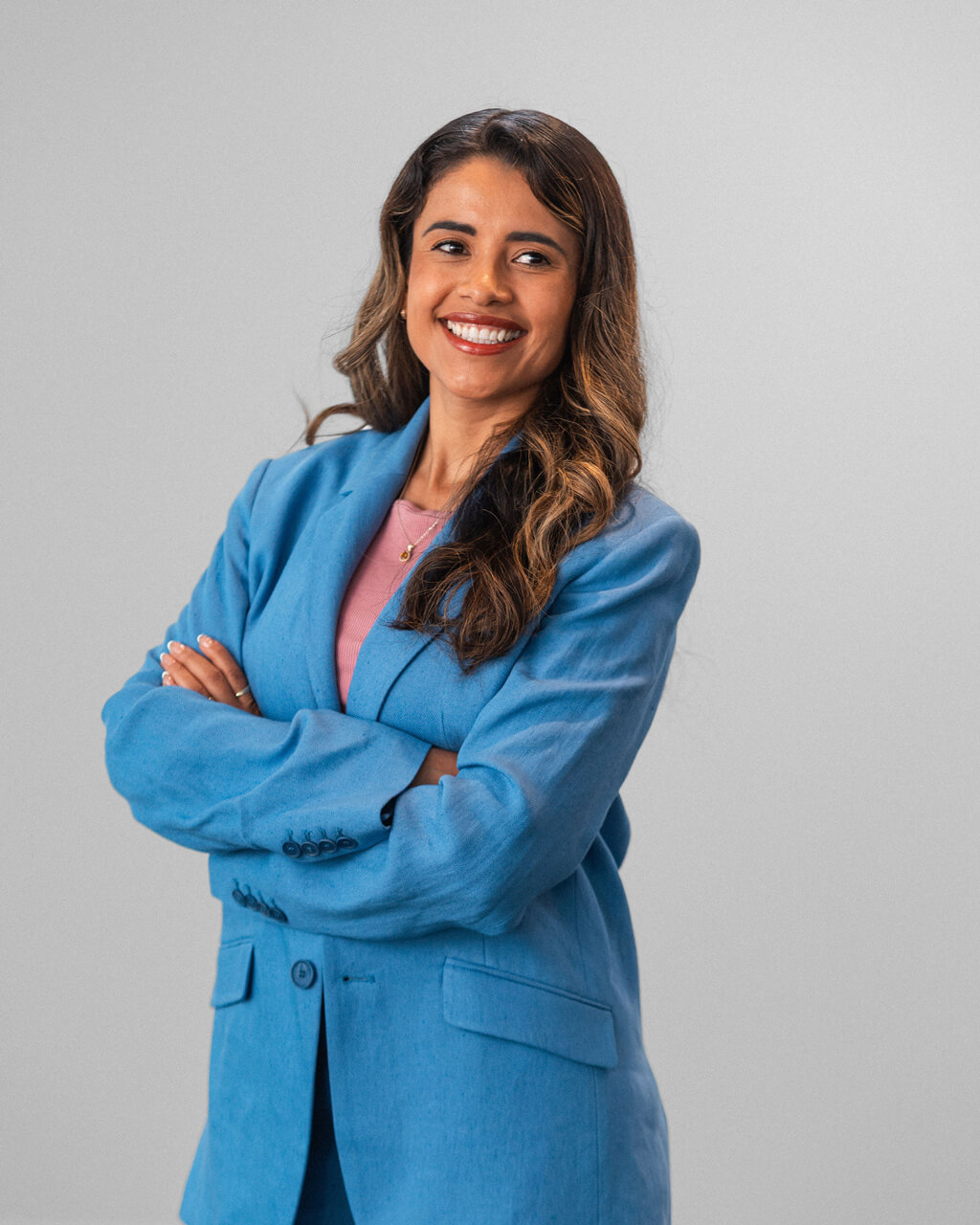Maira Jimenez, a woman with long brown hair, stands with arms crossed, smiling in a blue suit and pink shirt against a plain light gray background.