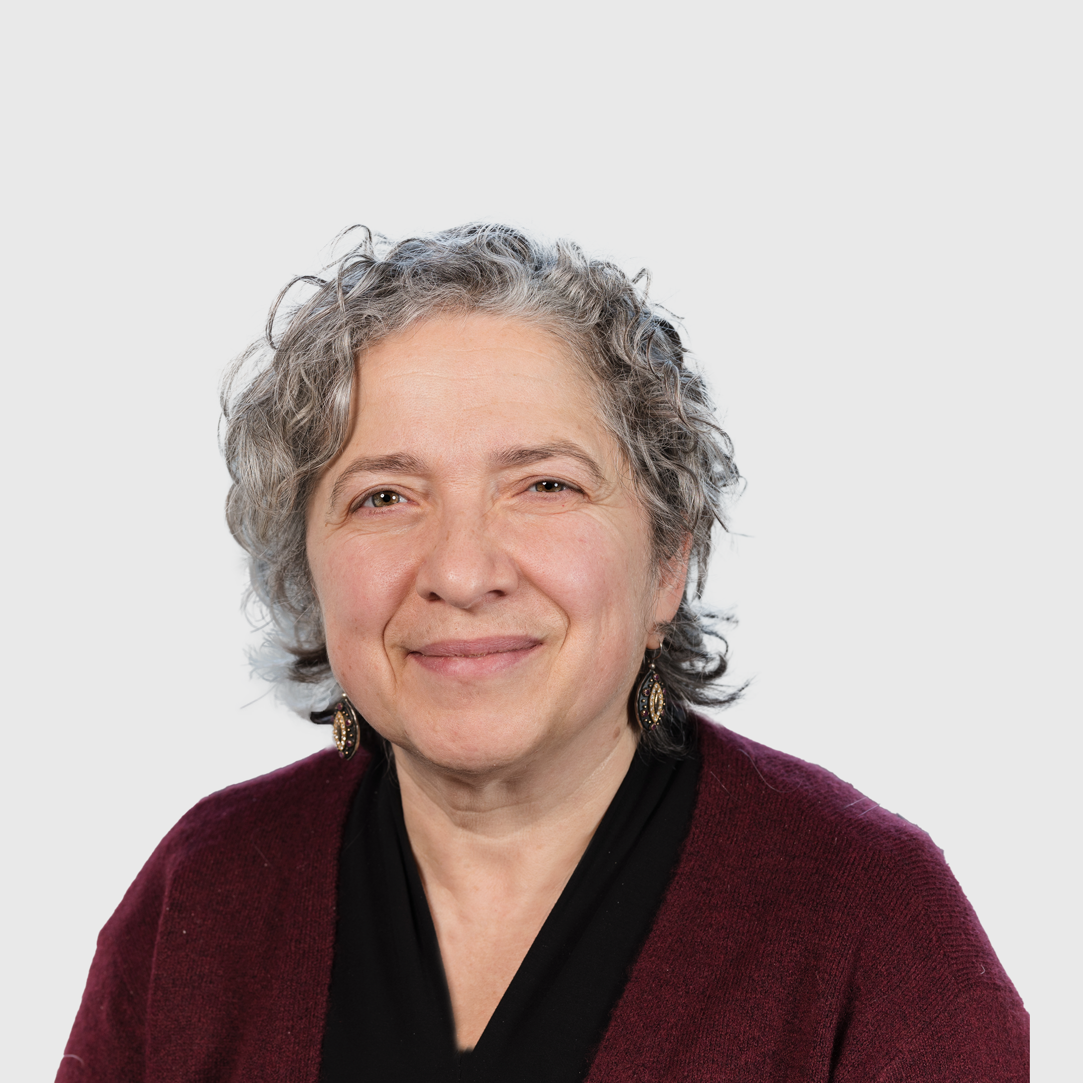Marina Bluvshtein, an older woman with short curly gray hair, wearing a maroon sweater, black top, and earrings, smiles at the camera against a plain light background.