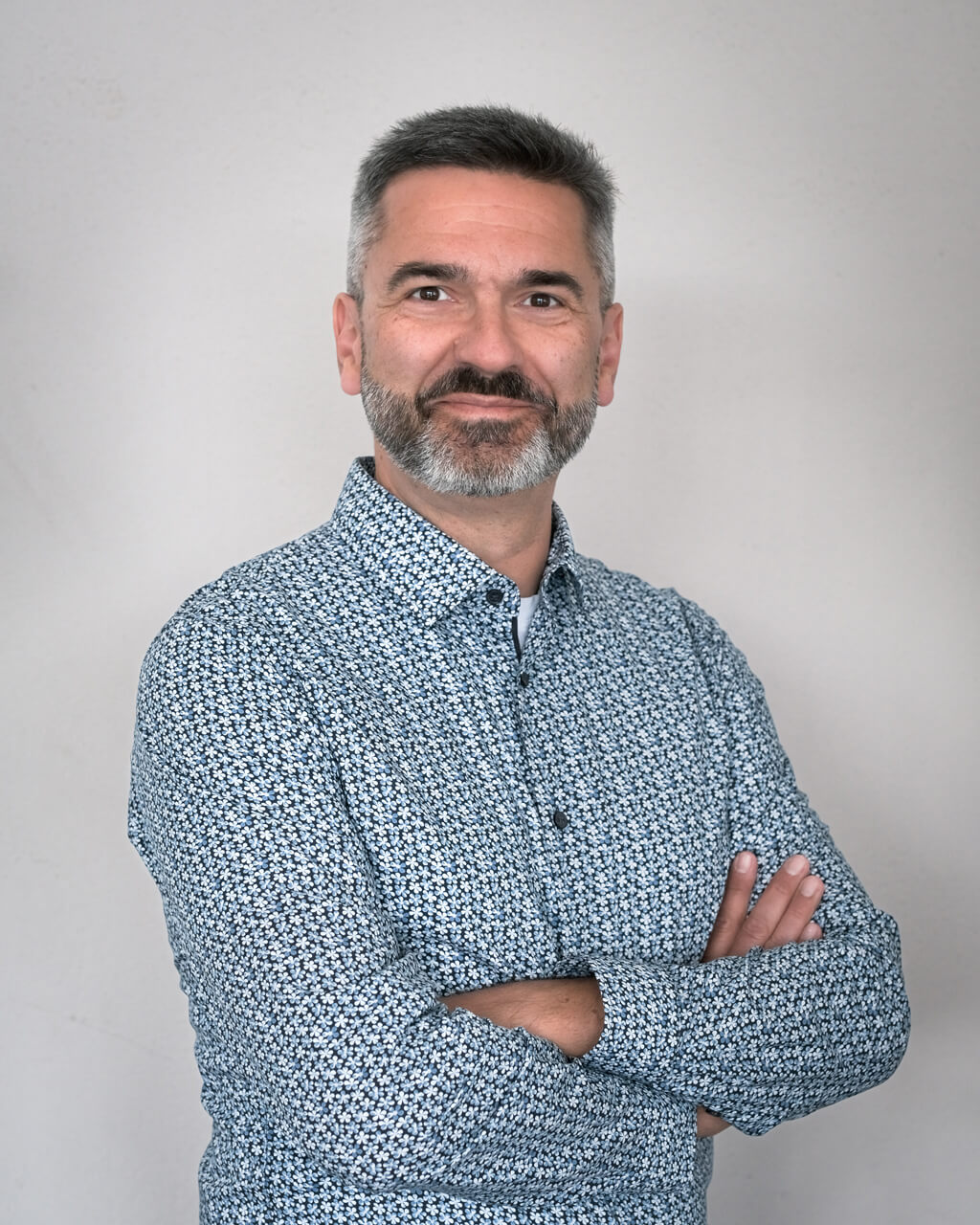 Michal Fedeles, a man with short gray hair and a beard, stands against a plain background, wearing a patterned button-up shirt and crossing his arms.