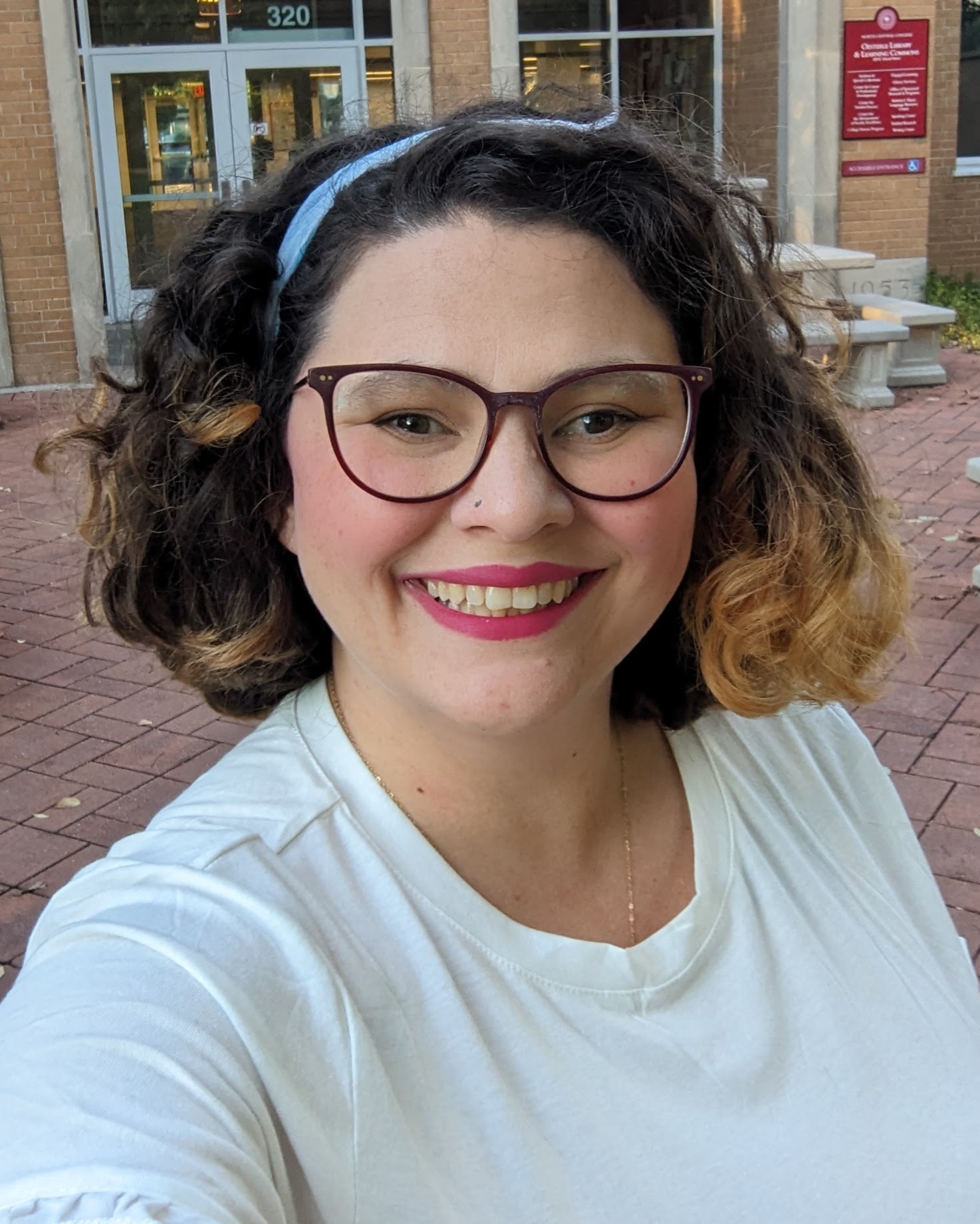 Cameron, with curly hair, glasses, and a blue headband, smiles while taking a selfie outdoors in front of a brick building.