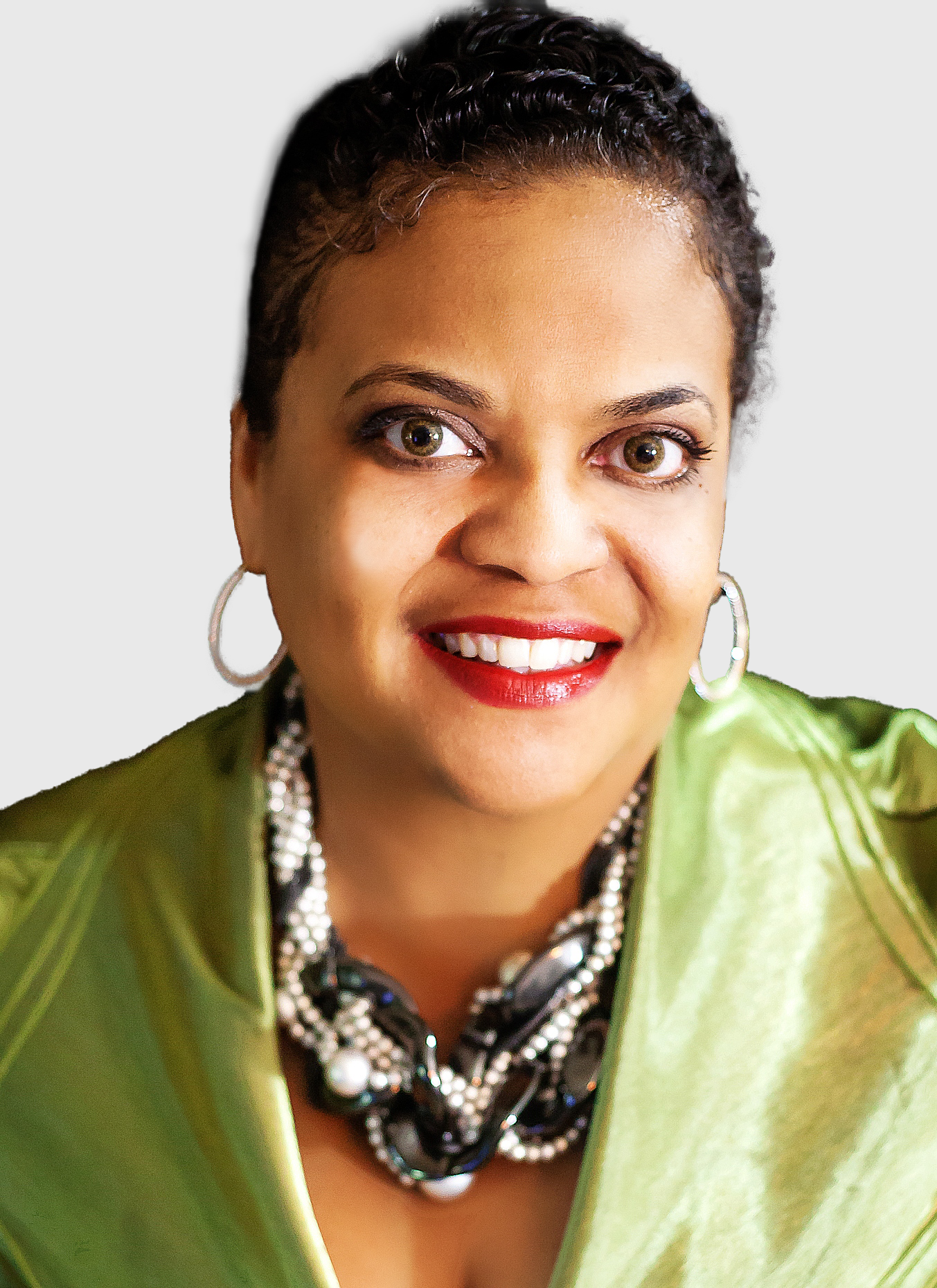 Phyllis Horton, a woman with short curly hair, wears a shiny green top, bold necklace, hoop earrings, and red lipstick as she smiles at the camera against a plain background.