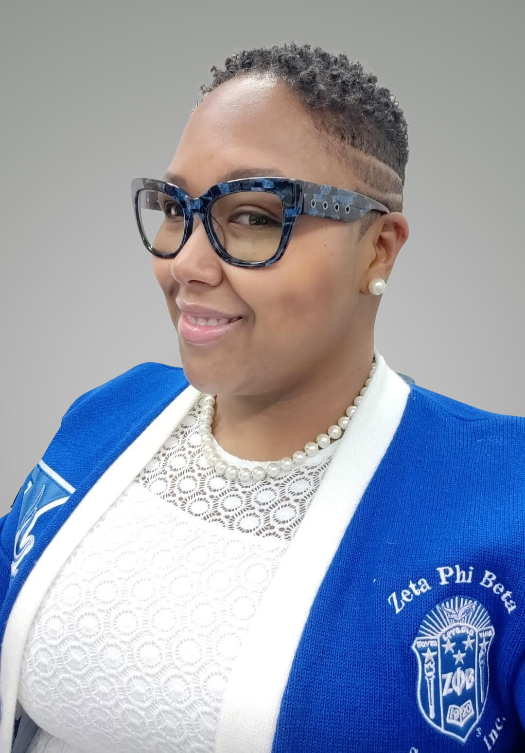 Shannon Thomison, a woman with short hair and glasses, wears a blue Zeta Phi Beta sorority cardigan, a white lace top, and a pearl necklace, smiling at the camera against a gray background.