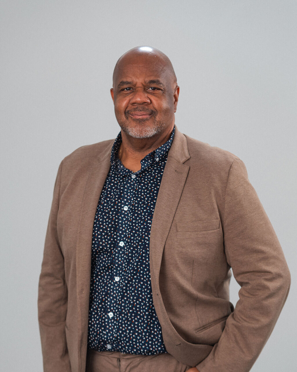 A middle-aged man with a bald head and gray beard—possibly Stephen S. Hinds—is wearing a tan blazer over a navy patterned shirt, standing against a plain light background.