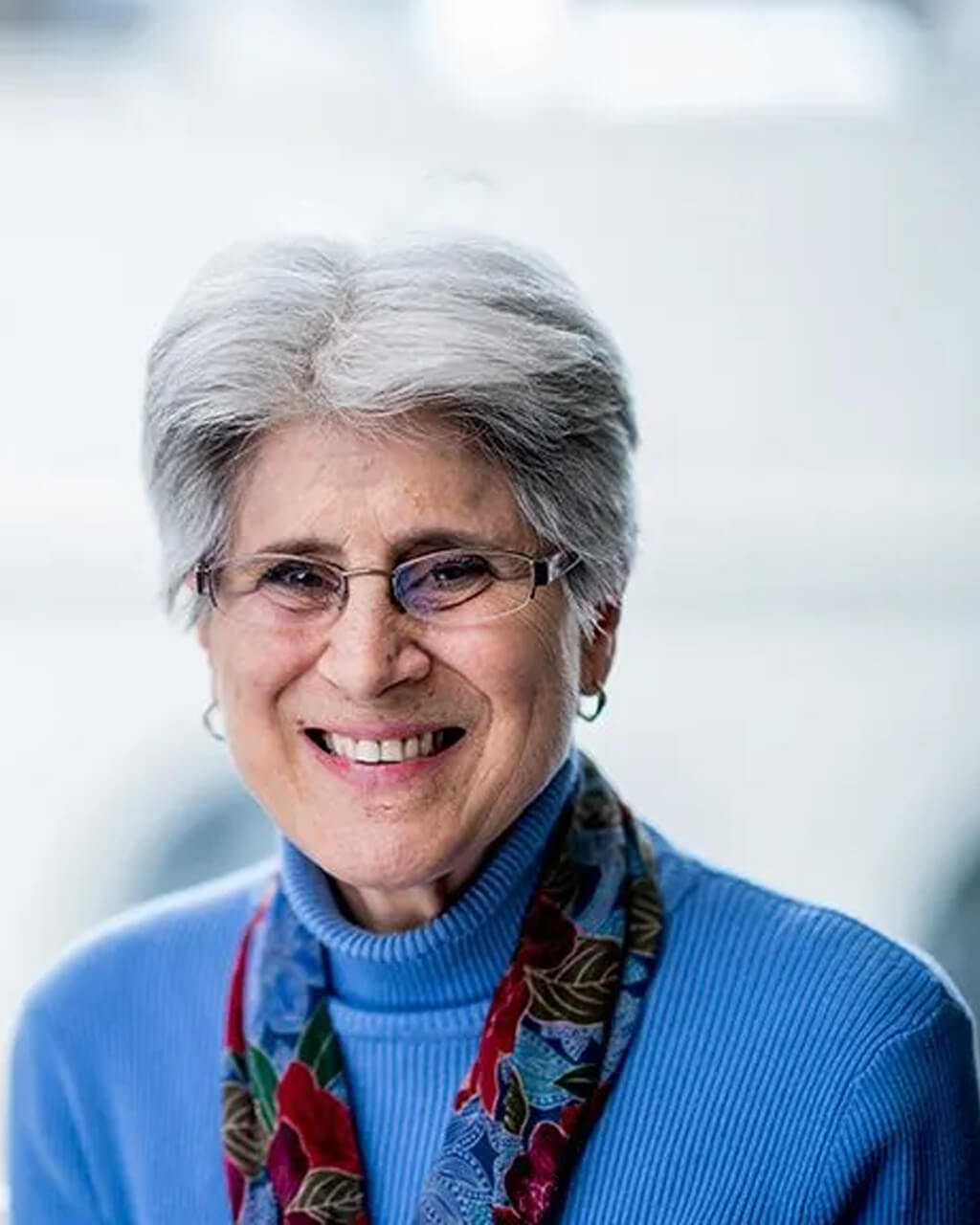 Susan S. Zoline, an older woman with short gray hair, eyeglasses, and a blue sweater with a floral scarf, smiles warmly at the camera against a blurred background.