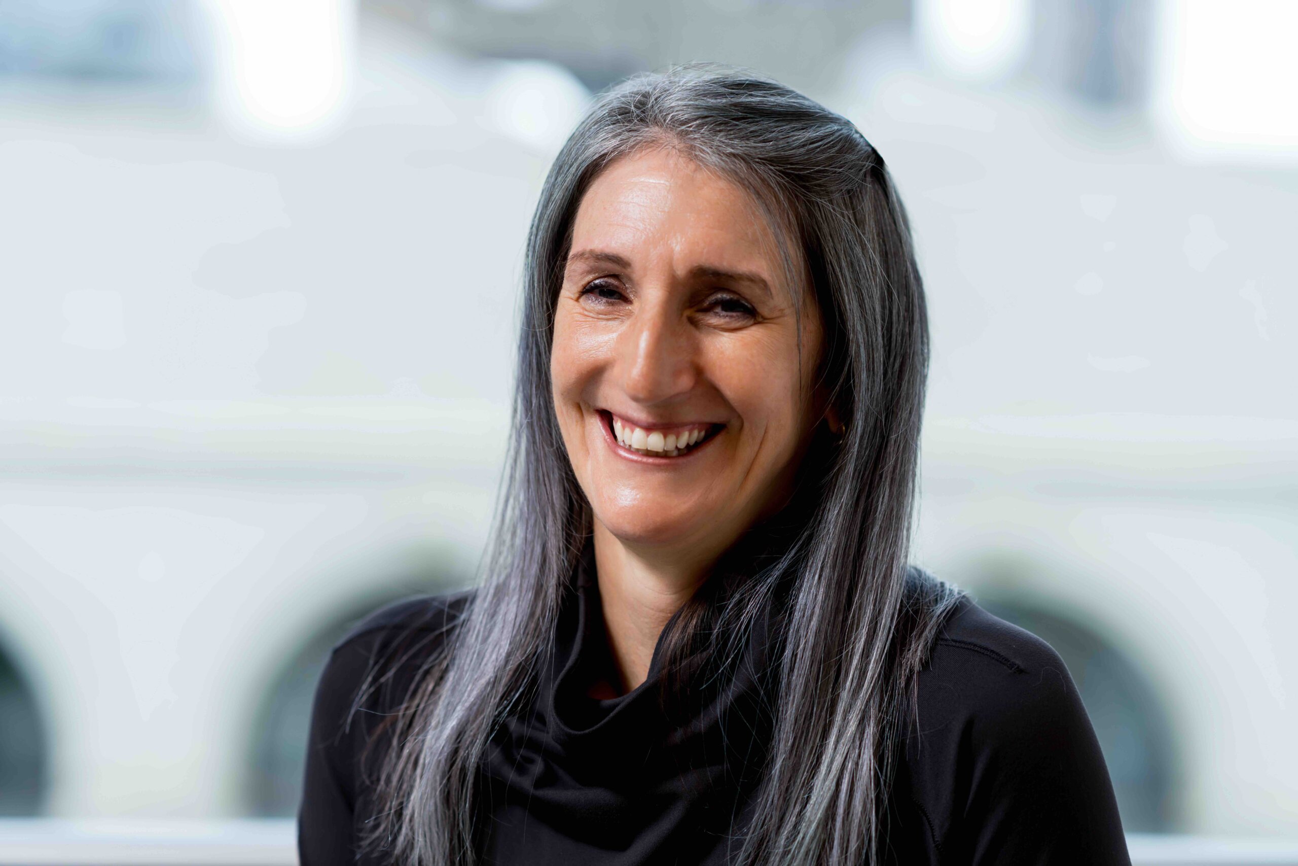 Teresa Behrend Fletcher, a woman with long gray hair and a black top, smiles at the camera, with a blurred background featuring white arches.
