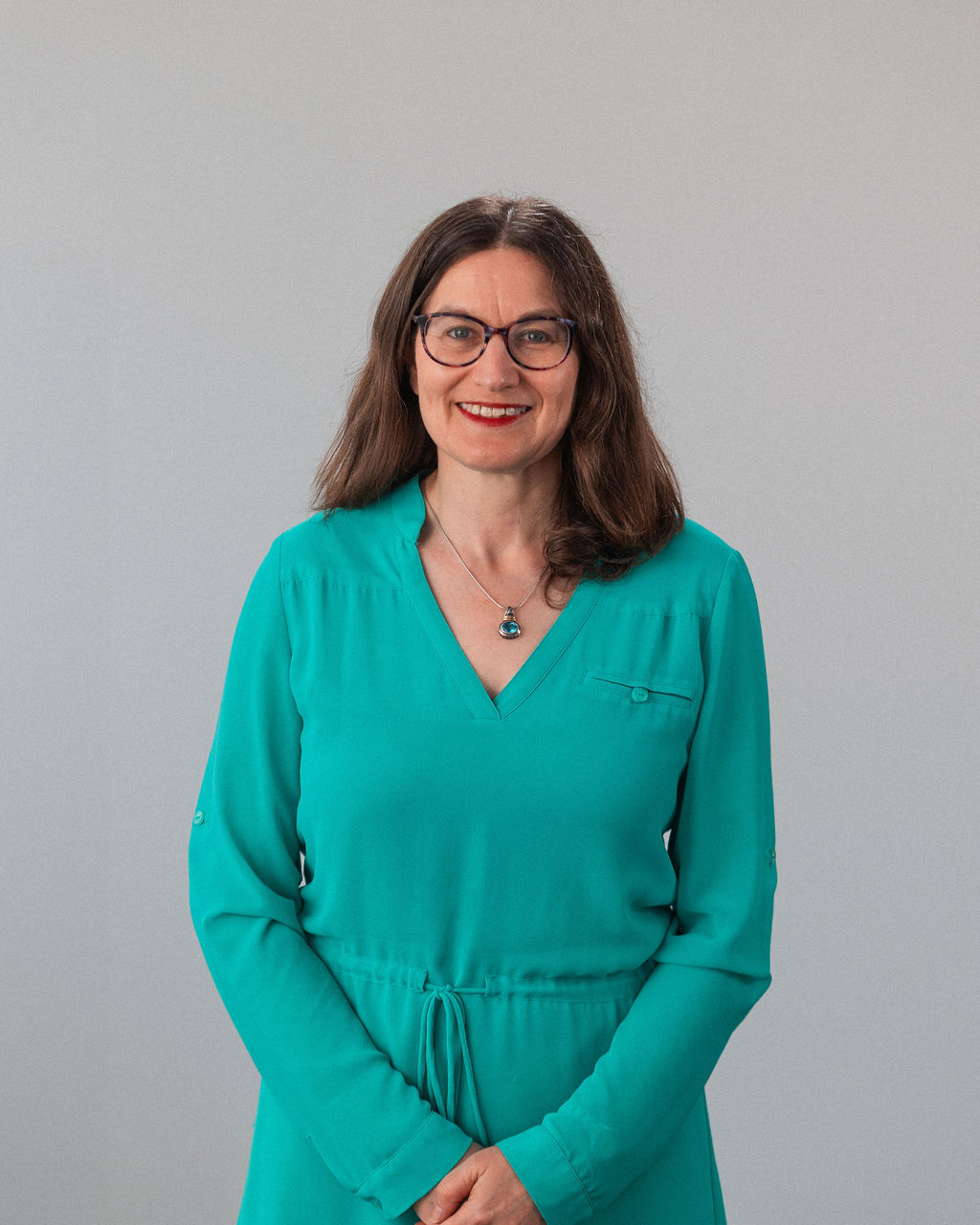 Nicole Dorfan, a woman with long brown hair, glasses, and a teal dress, stands against a plain light background, smiling with hands folded in front.