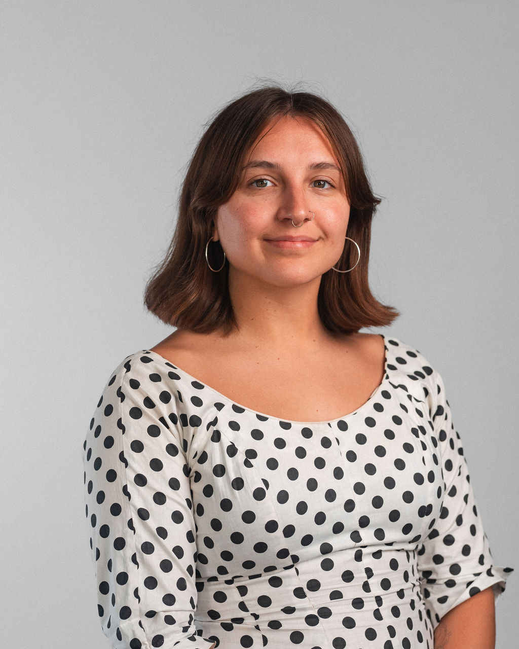 A woman with shoulder-length brown hair, wearing hoop earrings and a white dress with black polka dots, poses against a plain light gray background.