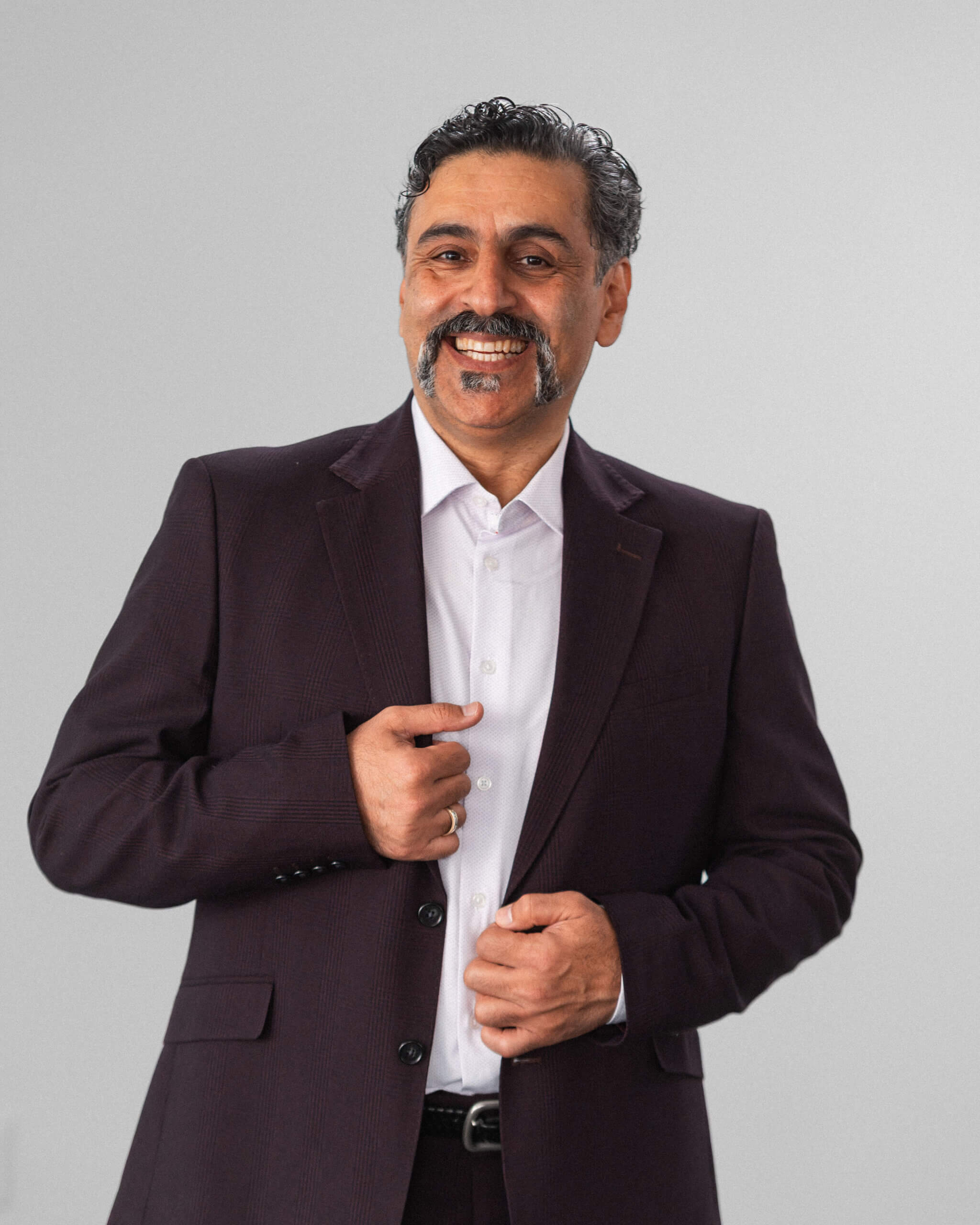 Alireza Sedghi Taromi, a man with dark hair and a mustache, stands against a plain light background in a dark suit and white shirt, smiling as he buttons his jacket.