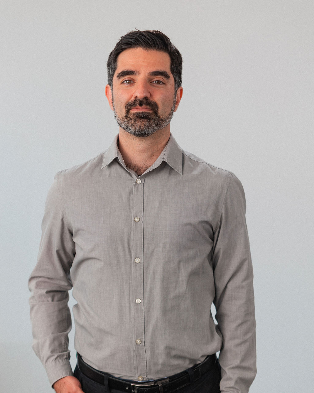 Babak, a man with short dark hair and a beard, wears a light gray button-up shirt and black pants while standing against a plain light gray background.