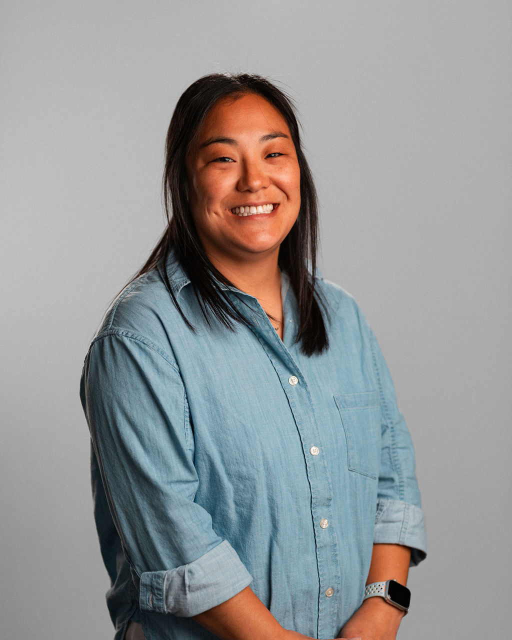 Terilyn C., with long dark hair and a light blue button-up shirt, stands smiling against a plain gray background, her smartwatch visible on her wrist.