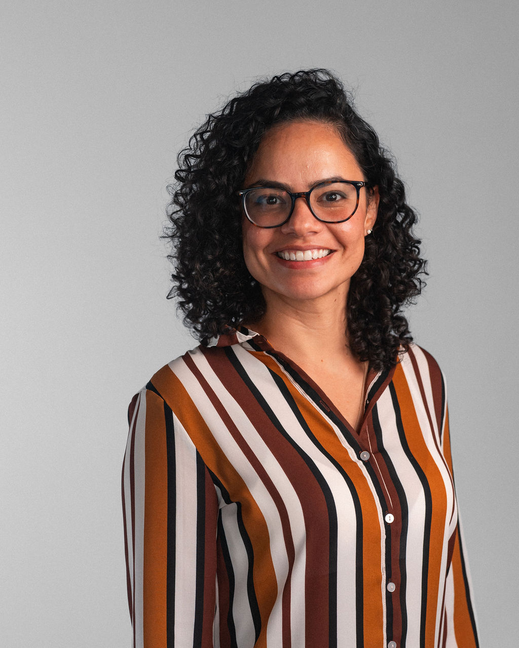 Reylla Santos, a woman with curly hair and glasses, smiles at the camera. She wears a button-up shirt with vertical brown, yellow, and white stripes, standing in front of a plain gray background.