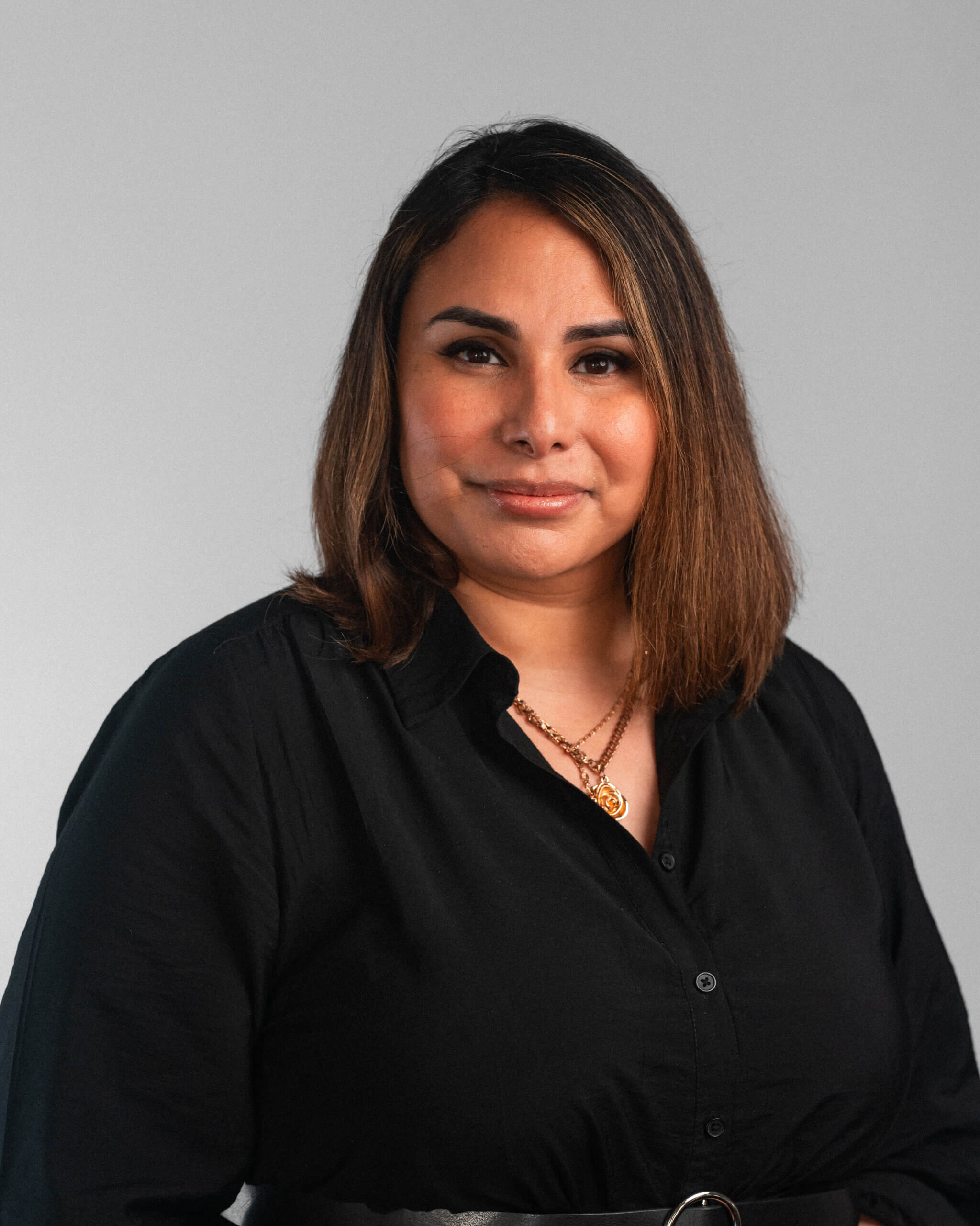 Karyna Strickland, a woman with straight, shoulder-length brown hair, wears a black button-up shirt and layered necklaces as she poses against a plain gray background.