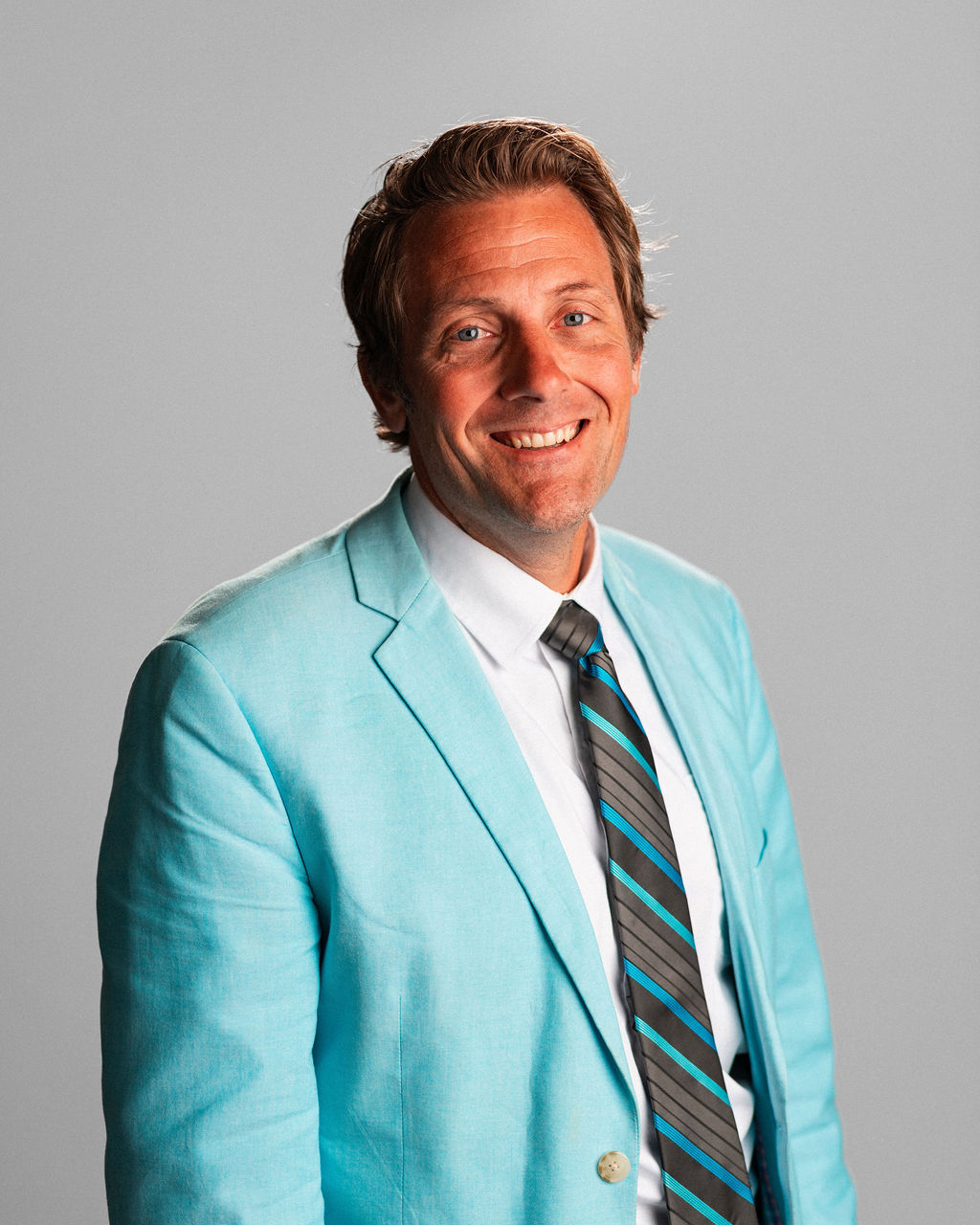 Ryan, wearing a light blue suit jacket, white shirt, and striped tie, stands smiling in front of a plain gray background.