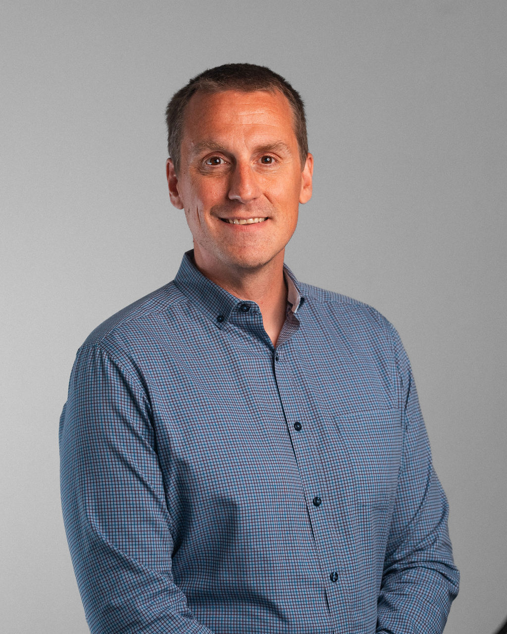 Eric J. Korslin, a man with short brown hair wearing a blue checkered button-up shirt, stands in front of a plain light gray background, facing the camera and smiling slightly.