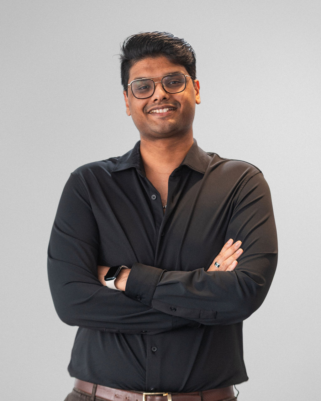 Kabir, wearing glasses and a black shirt, stands against a plain light background with his arms crossed, smiling at the camera.