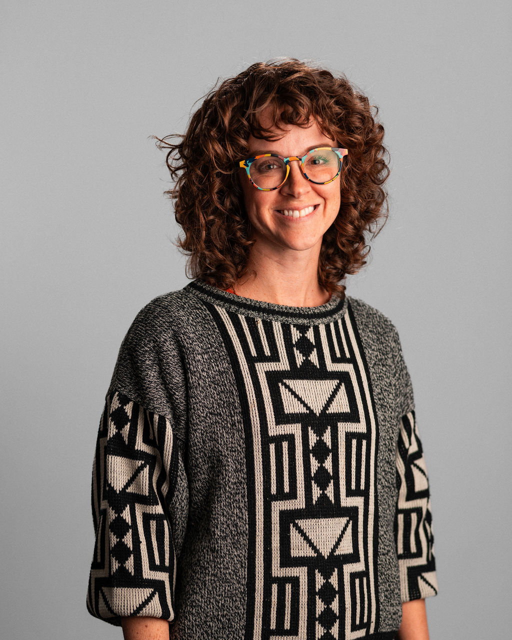Lauren Melamed, with curly brown hair, wearing glasses and a patterned sweater, stands smiling in front of a plain gray background.