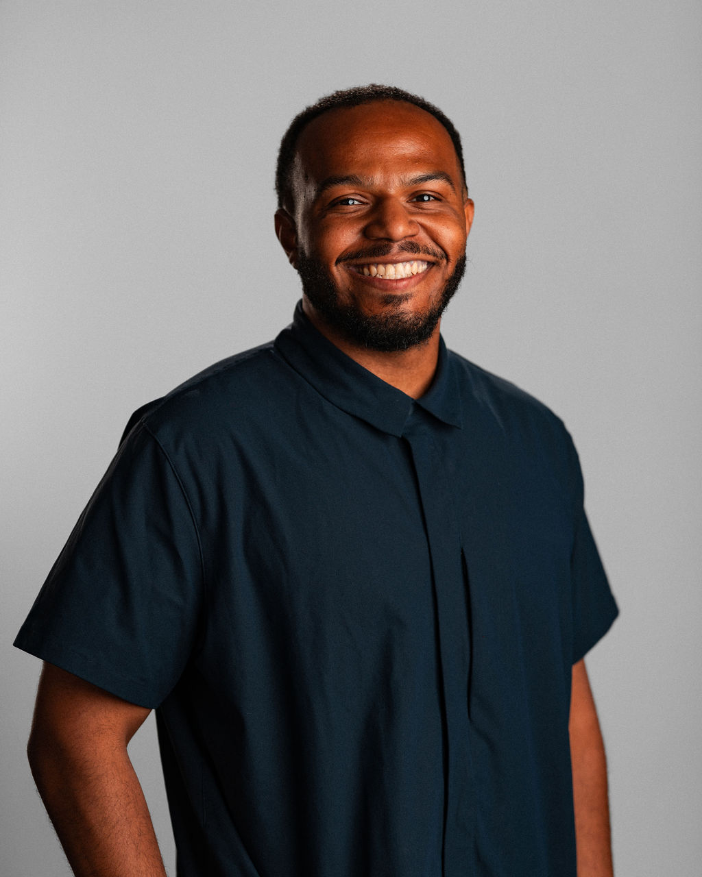 Brandon Huddleston, a man with short hair and a beard, is smiling while wearing a dark short-sleeve button-up shirt, standing against a plain light gray background.