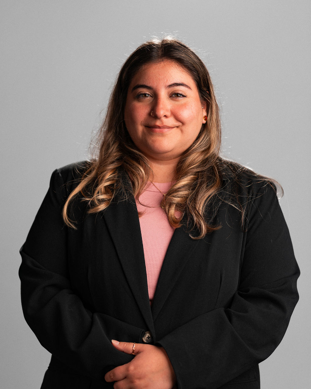 Izzy Peña Nakouzi, a woman with long, wavy hair, stands against a plain gray background in a black blazer over a pink top, facing the camera with a slight smile.