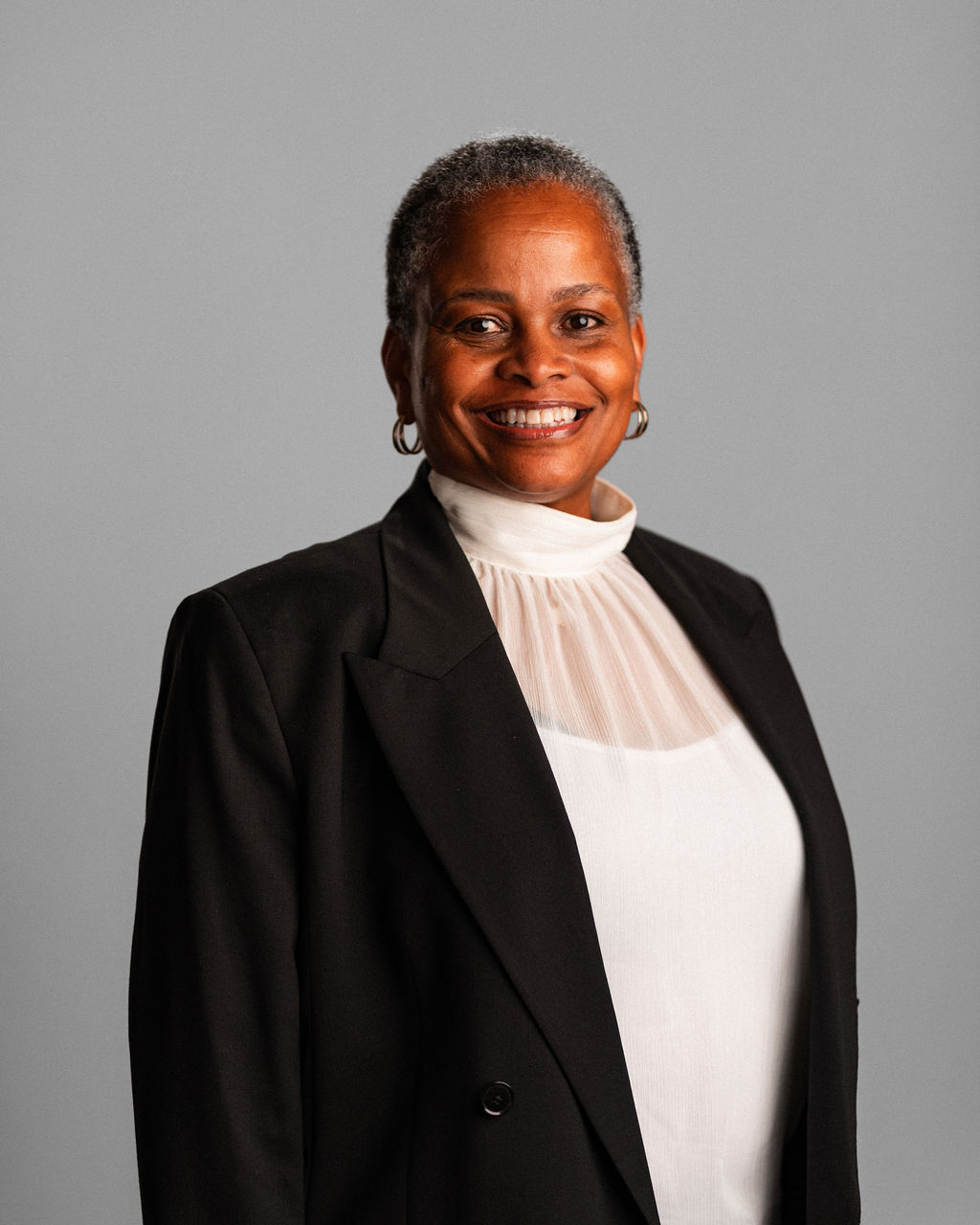 Cheryl Richardson, a woman with short gray hair, wearing a black blazer and white high-neck blouse, smiles at the camera against a plain gray background.
