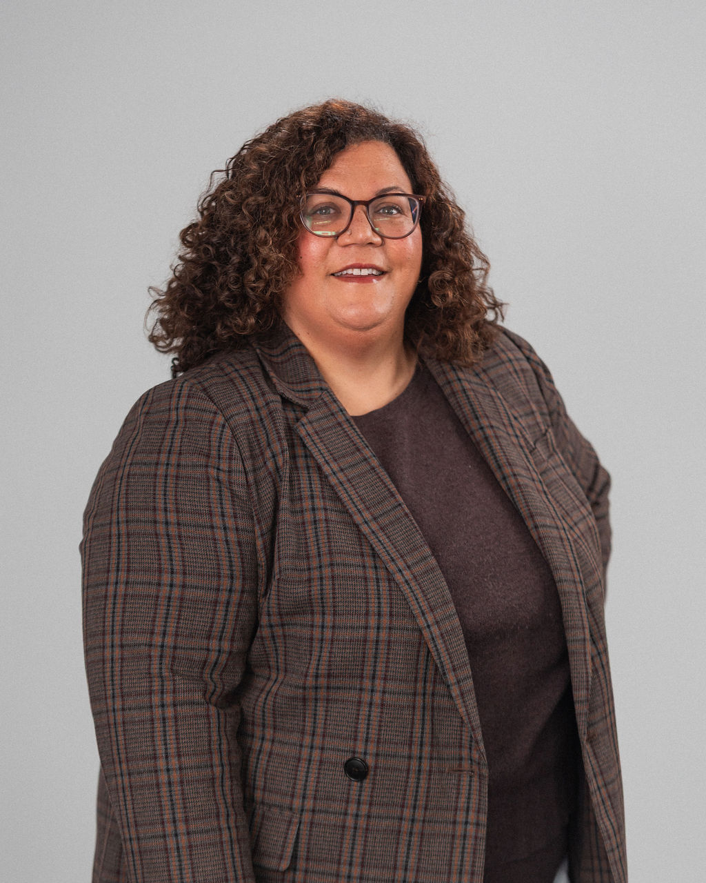 Susanne Milner, with curly hair and glasses, is wearing a plaid blazer and dark top, standing against a plain light gray background.