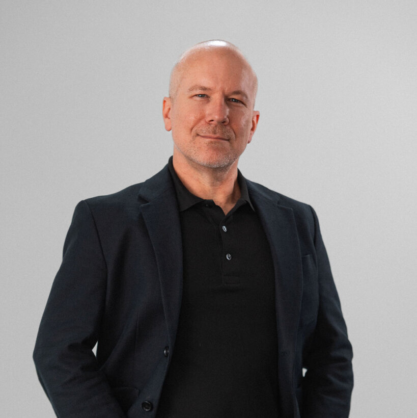 Aiken Benjamin, a middle-aged man with short gray hair, stands against a plain light gray background wearing a dark blazer over a black polo shirt.